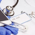Medizinisches Zubehör auf einer weißen Oberfläche, darunter ein Smartphone, ein Stethoskop, eine Namenskarte mit der Aufschrift "DR. med.", eine Brille, blaue Einweghandschuhe und medizinische Instrumente