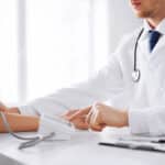 Ein Arzt misst den Blutdruck eines Patienten. Der Arzt trägt einen weißen Kittel und ein Stethoskop um den Hals, während er den Blutdruckmonitor überprüft.